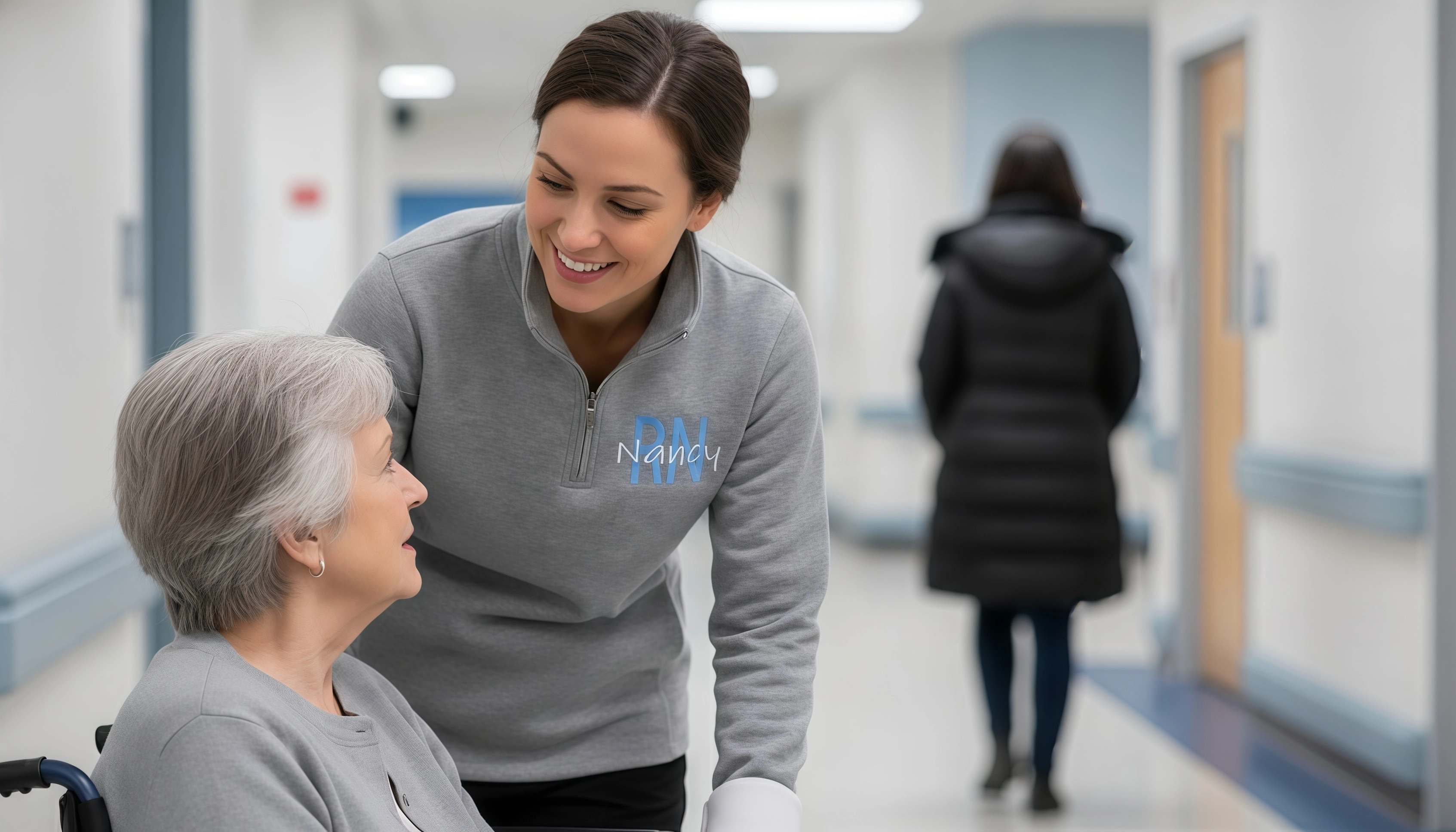 Registered nurse wearing a lightweight personalized RN jacket with custom embroidery, listening to a patient in a wheelchair in a hospital corridor.